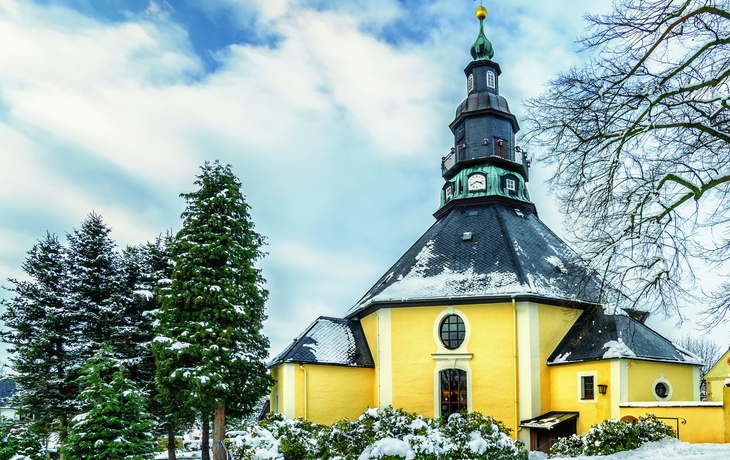 Gelbe Kirche im Schnee mit blauem Himmel und Tannenbäumen.