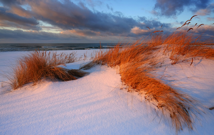Kolobrzeg im Winter© Konrad Uznański - stock.adobe.com Schneebedeckte Dünen am Meer bei Sonnenaufgang mit Wolken am Himmel.