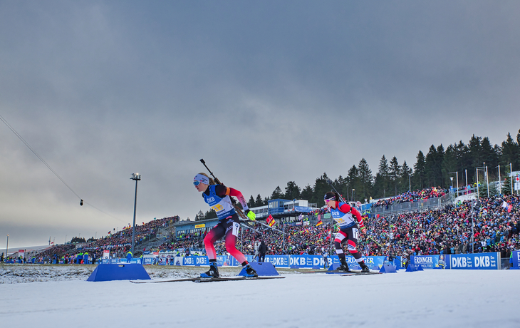 Zwei Biathleten beim Weltcup-Rennen auf schneebedeckter Strecke mit Publikum.
