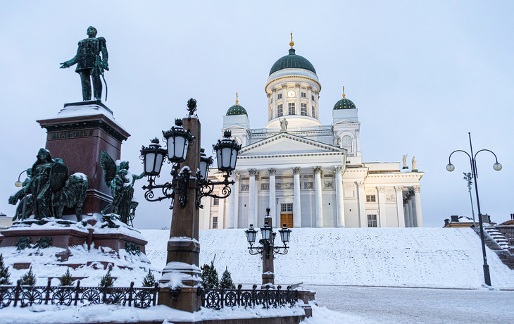 Panoramablick auf die Kathedrale von Helsinki im Winter bei Dämmerung, umgeben von Schnee und einer Statue im Vordergrund.