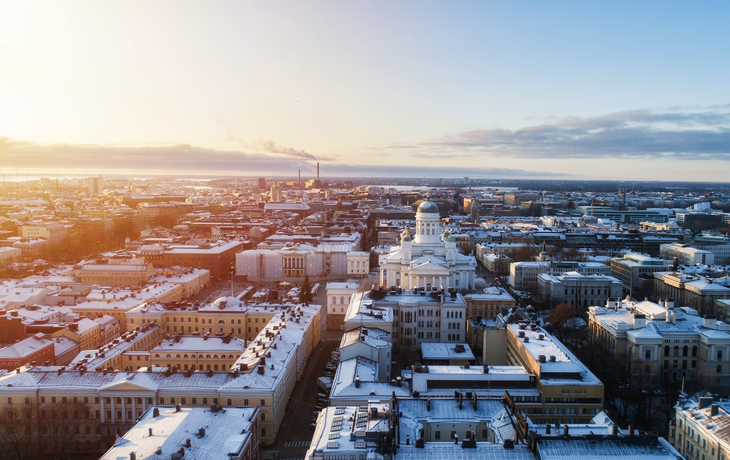 Luftaufnahme von Helsinki im Winter, mit schneebedeckten Gebäuden und der weißen Kuppel der Kathedrale im Zentrum, umgeben von einer städtischen Landschaft unter einem klaren blauen Himmel.
