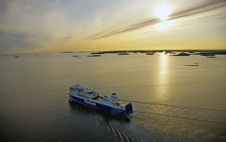 Ein großes Fährschiff der Reederei Finnlines fährt bei Sonnenuntergang durch offene Gewässer, im Hintergrund sind mehrere kleine Inseln und die untergehende Sonne zu sehen.