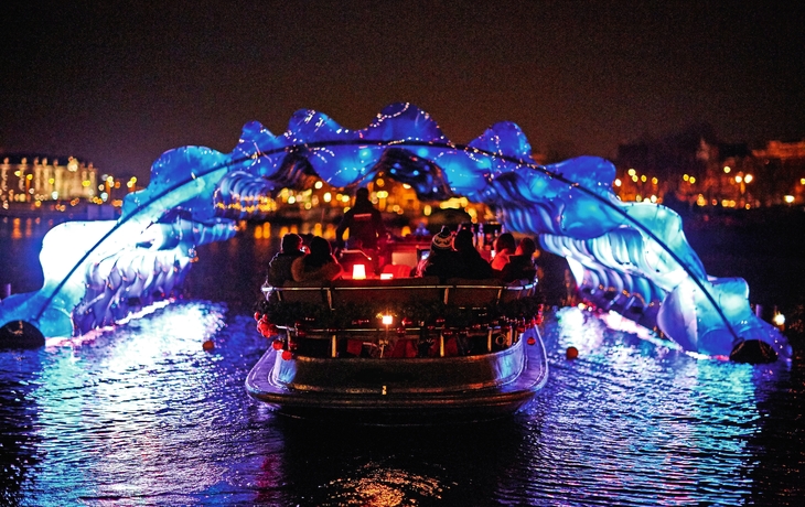 Boot fährt nachts unter blau beleuchtetem, wellenförmigem Kunstwerk auf dem Wasser.