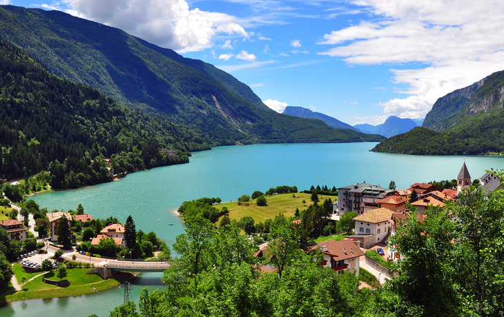 Blick auf einen See mit umliegenden Bergen und einem Dorf im Vordergrund.