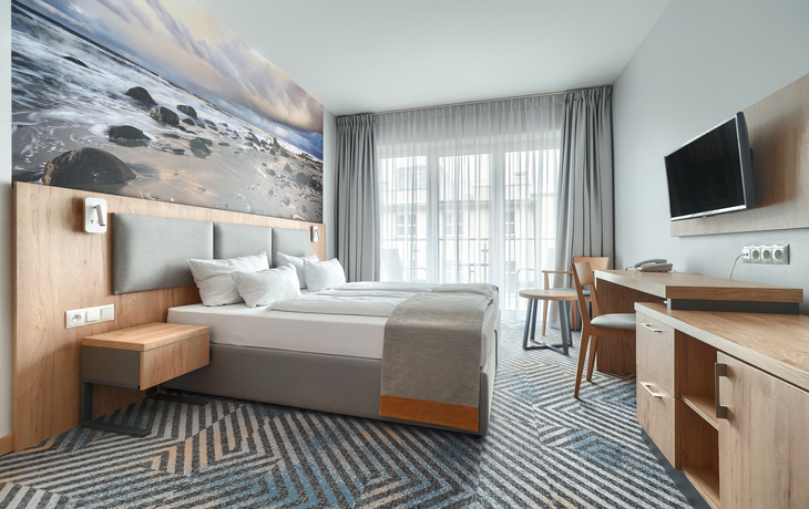 Modernes Hotelzimmer mit Doppelbett, Schreibtisch, Fernseher und großem Fenster.