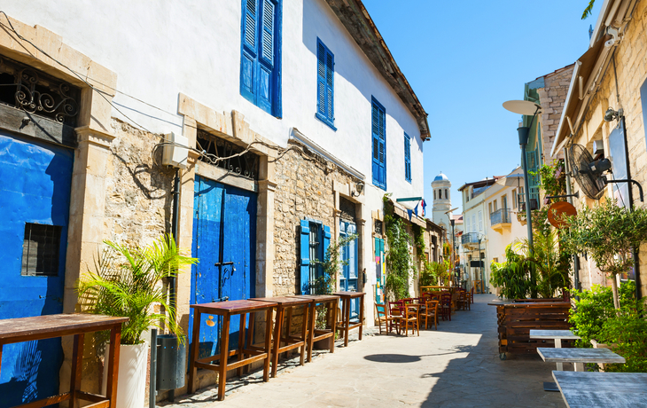 Gasse mit historischen Gebäuden, blauen Fensterläden und Außenterrasse bei Sonnenschein.