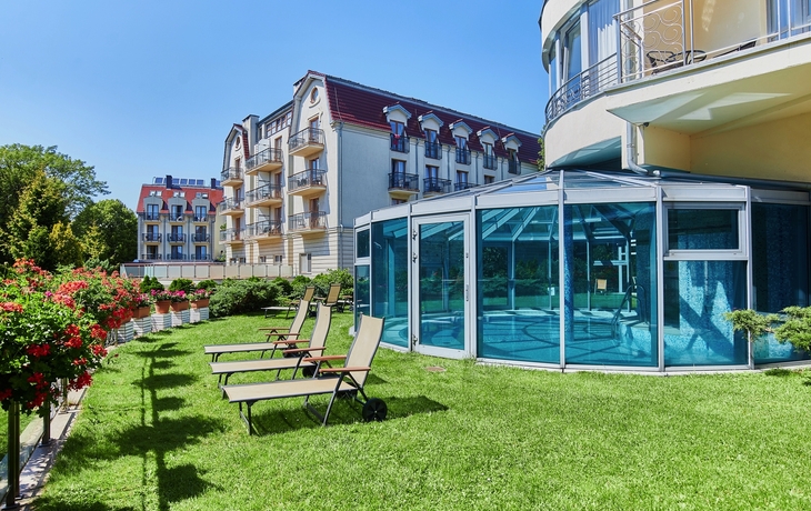 Gebäude mit Balkon und verglastem Schwimmbad neben Liegestühlen auf Rasenfläche.