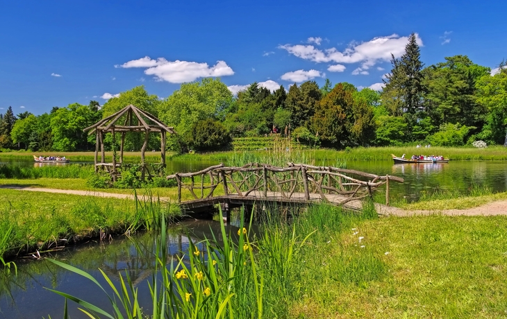 Grüne Parklandschaft mit Holzbrücke, Pavillon und Booten im Wasser.