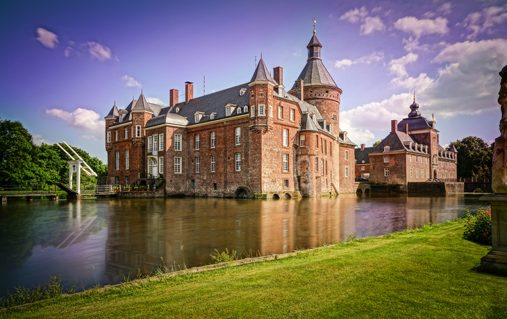 Backsteinschloss mit Türmen am Wasser, umgeben von grüner Landschaft.