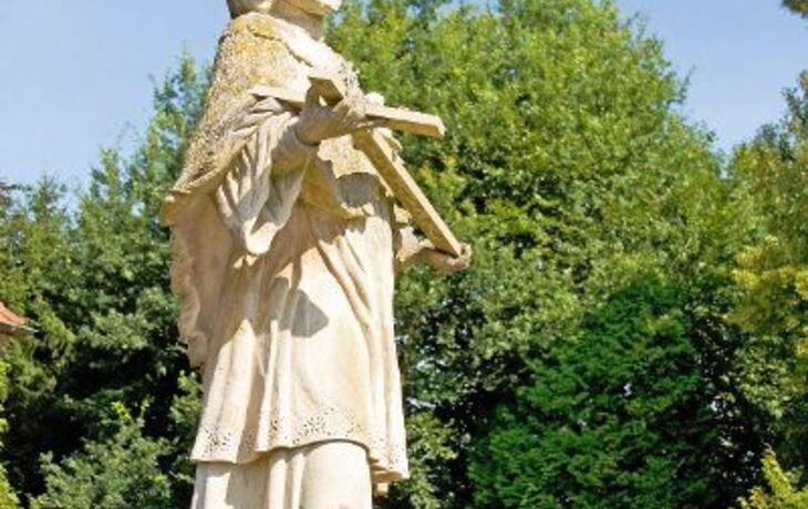 Steinskulptur eines Mannes mit Krone und Buch vor Bäumen im Freien.