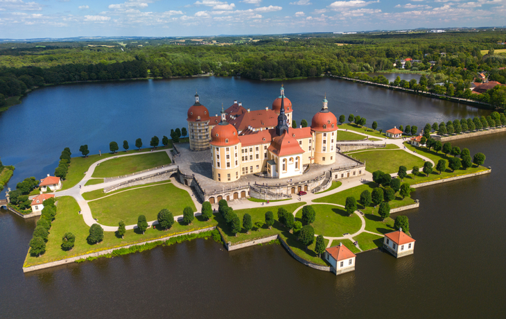Barockschloss mit roten Dächern auf Insel in malerischer Landschaft mit Wasser und Wald.