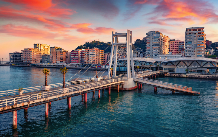 Hängebrücke über Wasser mit Stadt im Hintergrund bei Sonnenuntergang.