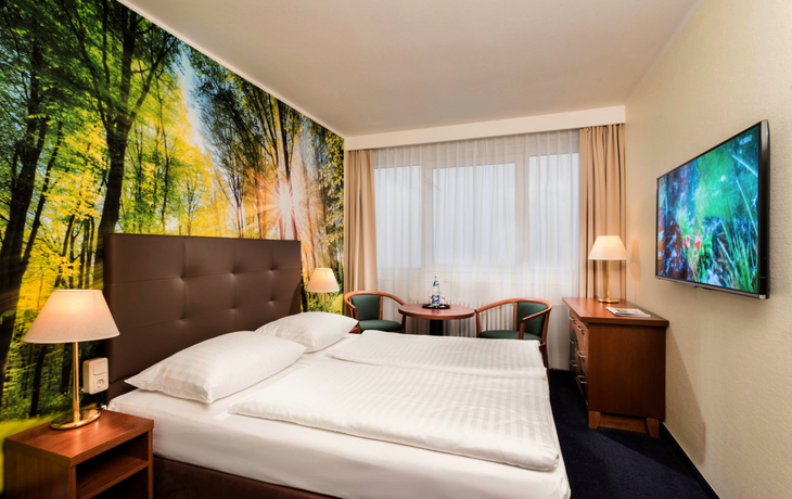 Hotelzimmer mit Doppelbett, Waldbild an der Wand, Fenster, TV, Tisch und Stühle.