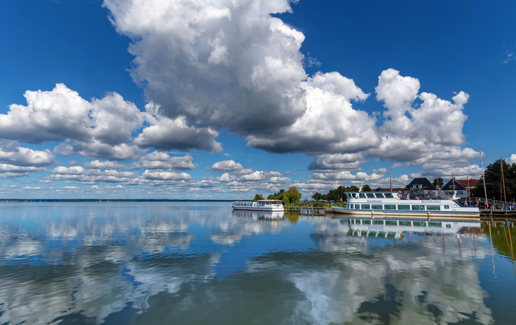 Wolken spiegeln sich in einem ruhigen See, zwei Boote am Ufer, blauer Himmel.