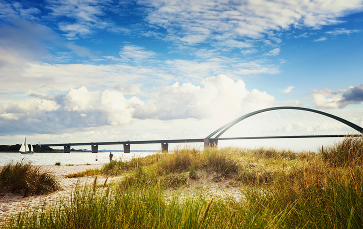 Sandstrand mit Dünengras, im Hintergrund eine Brücke über das Meer bei sonnigem Wetter.