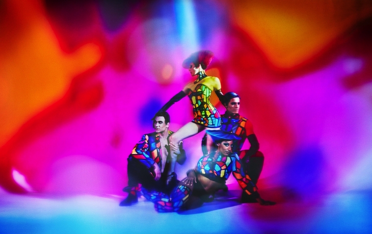 Vier Personen in bunten, geometrischen Kostümen vor lebhaftem, verschwommenem Hintergrund.