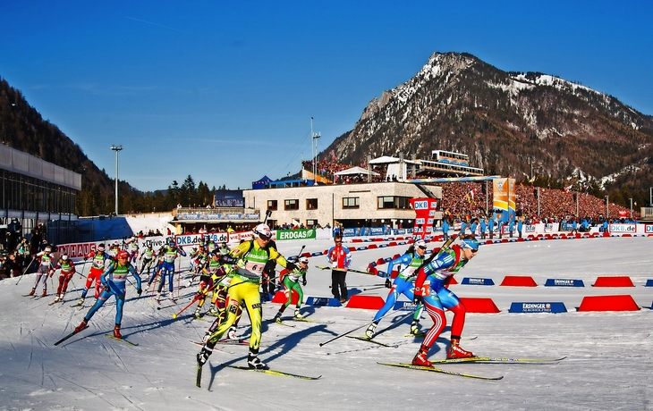 Biathleten beim Wettkampf in der Chiemgau Arena mit Bergen im Hintergrund.
