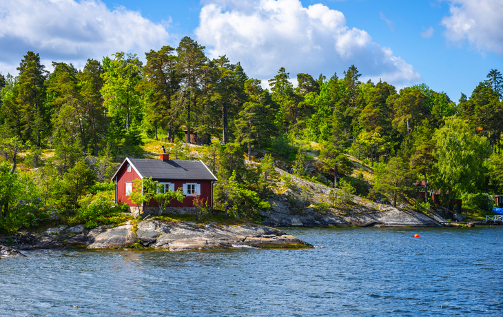 Rotes Häuschen am Seeufer, umgeben von Wald und blauem Himmel.