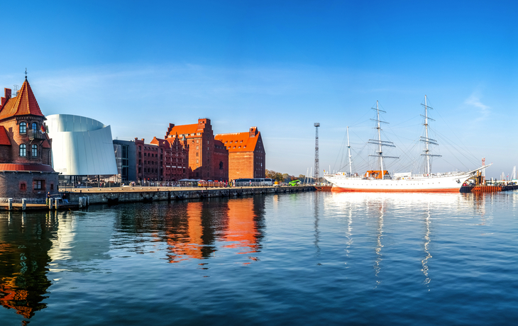 Panorama einer Hafenstadt mit historischen Gebäuden und einem Segelschiff im Wasser.