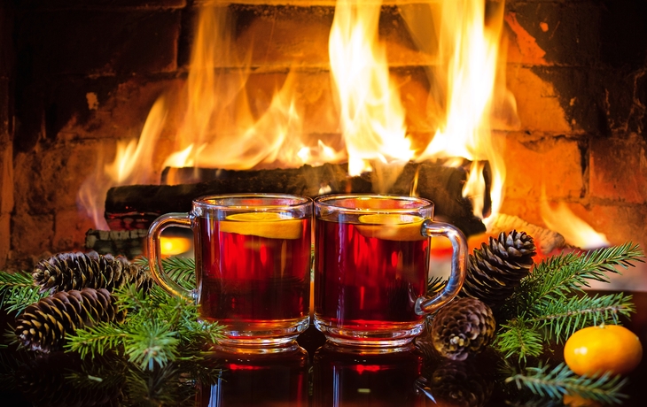 Zwei Glühweingläser vor einem Kamin mit Tannenzapfen und Weihnachtsdeko.
