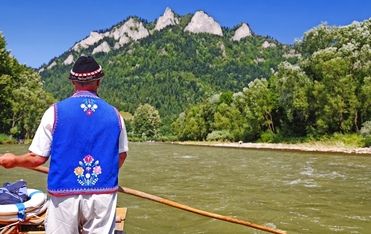 Ein Mann in traditioneller Kleidung steuert ein Boot auf einem Fluss vor bergiger Landschaft.