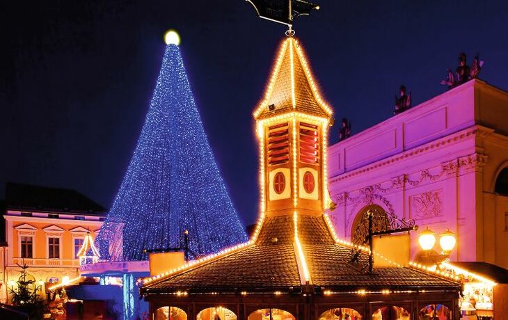 Beleuchteter Weihnachtsmarktstand vor dekoriertem Baum bei Nacht.