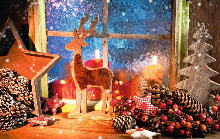 Weihnachtliche Dekoration mit Kerzen, Rentierfigur und Tannenzapfen vor einem Fenster.