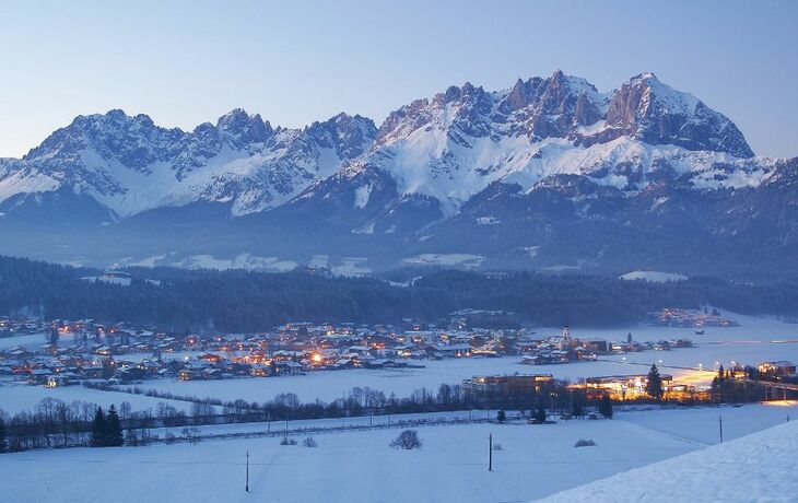 Winterliches Dorf vor verschneiten Bergen und beleuchteten Häusern in der Dämmerung.