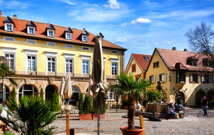 Historischer Platz mit gelben Gebäuden, Sonnenschirmen und Palmen bei klarem Himmel.