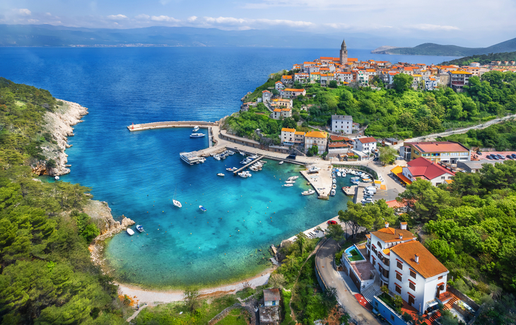 Küstenstadt mit Bucht, Booten und farbenfrohen Gebäuden auf Hügel in sonniger Landschaft.