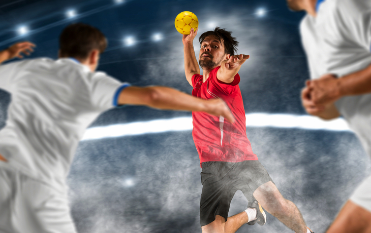 Handballspieler in rotem Trikot wirft Ball während eines Spiels in der Halle.