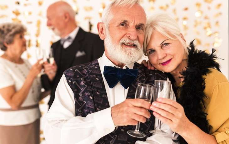 Älteres Paar in festlicher Kleidung stößt mit Sektgläsern an, im Hintergrund weiteres Paar.