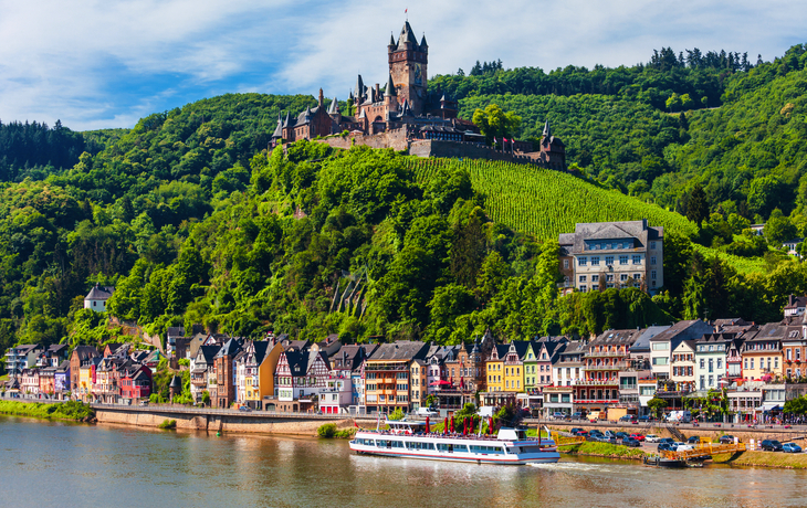 Reichsburg Cochem überragt Moselpromenade mit Schiffen im Vordergrund.