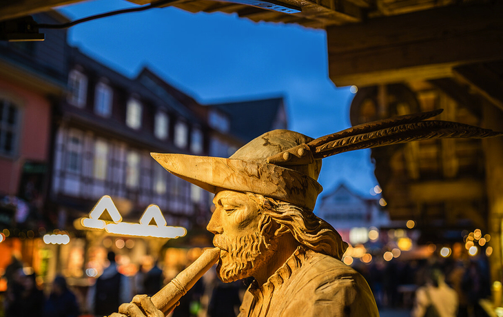 Skulptur eines Mannes mit Hut und Flöte auf einem belebten Markt bei Abenddämmerung.