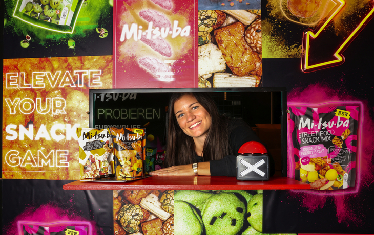 Frau präsentiert Snacks vor farbenfrohem Werbeaufsteller.