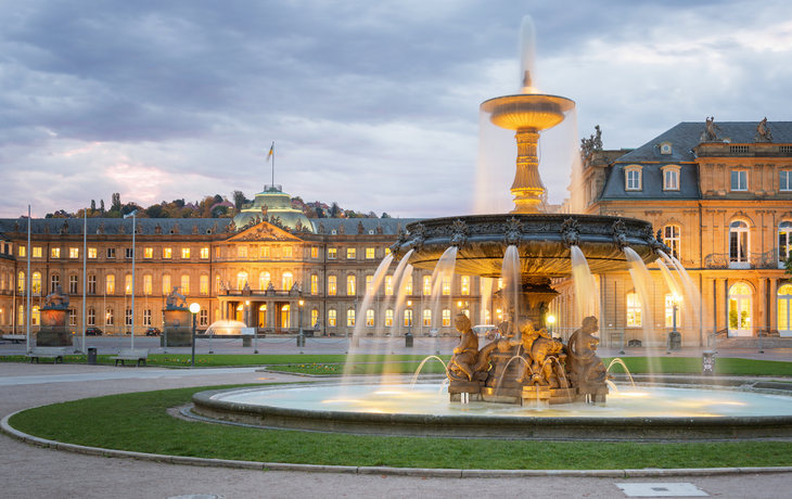 Brunnen vor einem beleuchteten Schloss bei bewölktem Himmel.