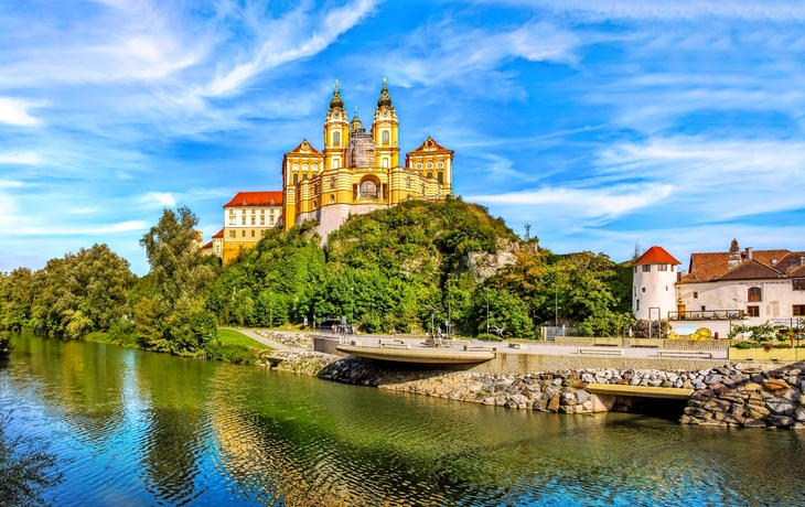 Barockes Kloster auf Hügel über Fluss, umgeben von Bäumen und blauem Himmel.