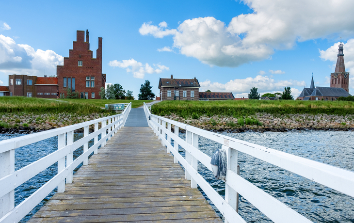 Holzsteg führt über Wasser zu Gebäuden unter blauem Himmel.