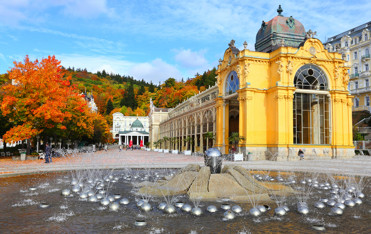 Hübscher historischer Brunnen vor einem gelben Kurhaus mit herbstlicher Landschaft.