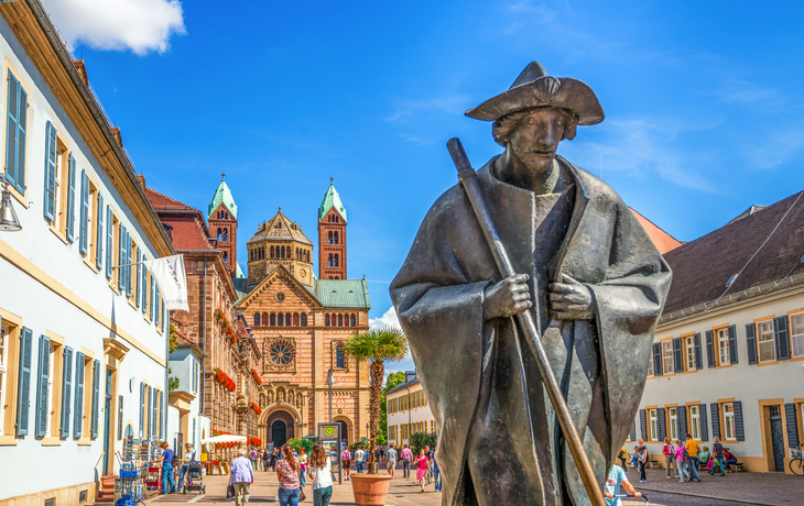 Statue eines Pilgers vor dem Speyerer Dom in einer belebten Fußgängerzone.