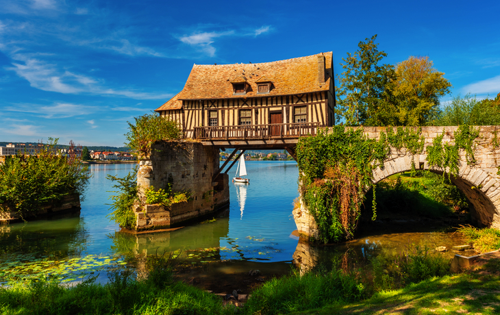 Fachwerkhaus auf einer alten Steinbrücke über einem Fluss mit Segelboot im Hintergrund.