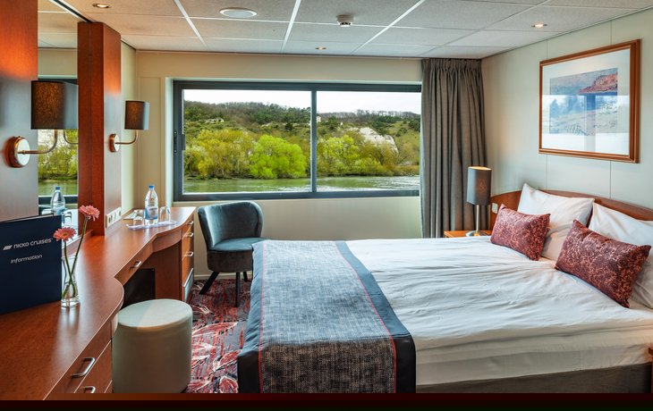 Kabine mit Doppelbett, Schreibtisch und großem Fenster mit Aussicht auf Flusslandschaft.