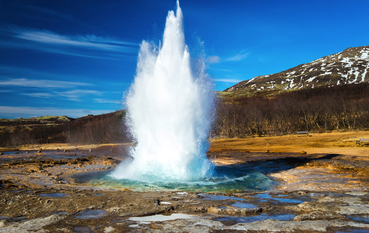 Geysir sprüht Wasser in die Luft vor Berge und blauem Himmel.