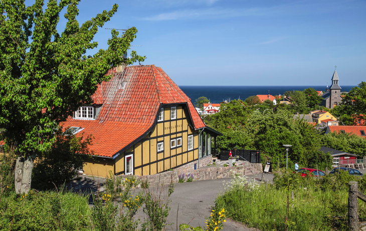 Fachwerkhaus mit rotem Dach in grüner Landschaft, im Hintergrund Meer und Kirchturm.