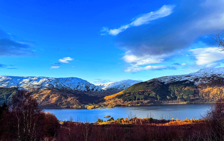 Berglandschaft mit See, bewaldeten Hängen und teilweise schneebedeckten Gipfeln.