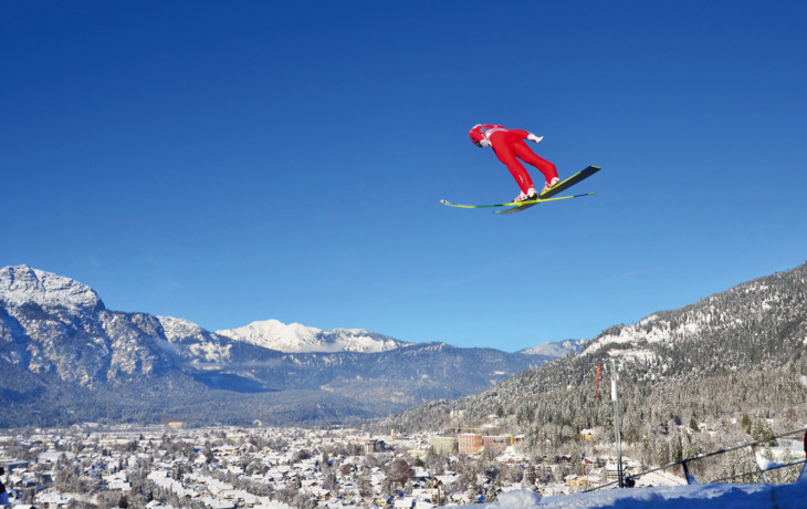 Skispringer in rotem Anzug fliegt über schneebedeckte Stadt und Berglandschaft.