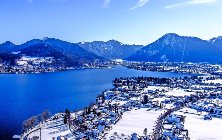Verschneite Stadt am Ufer eines Sees mit Bergen im Hintergrund.