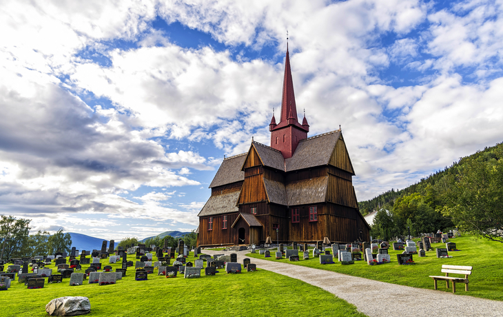 Holzkirche mit Friedhof, umgeben von grüner Landschaft unter bewölktem Himmel.