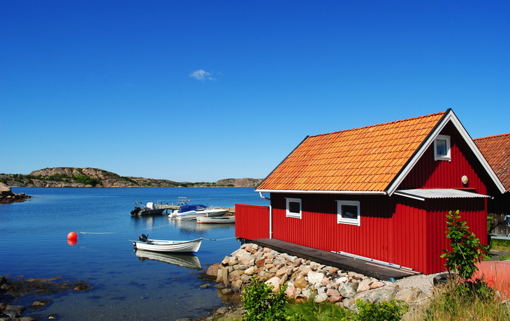 Rot gestrichenes Haus am Wasser mit Booten und blauem Himmel.