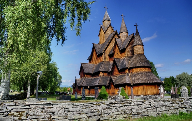Holzstabkirche mit Friedhof und Bäumen bei blauem Himmel.